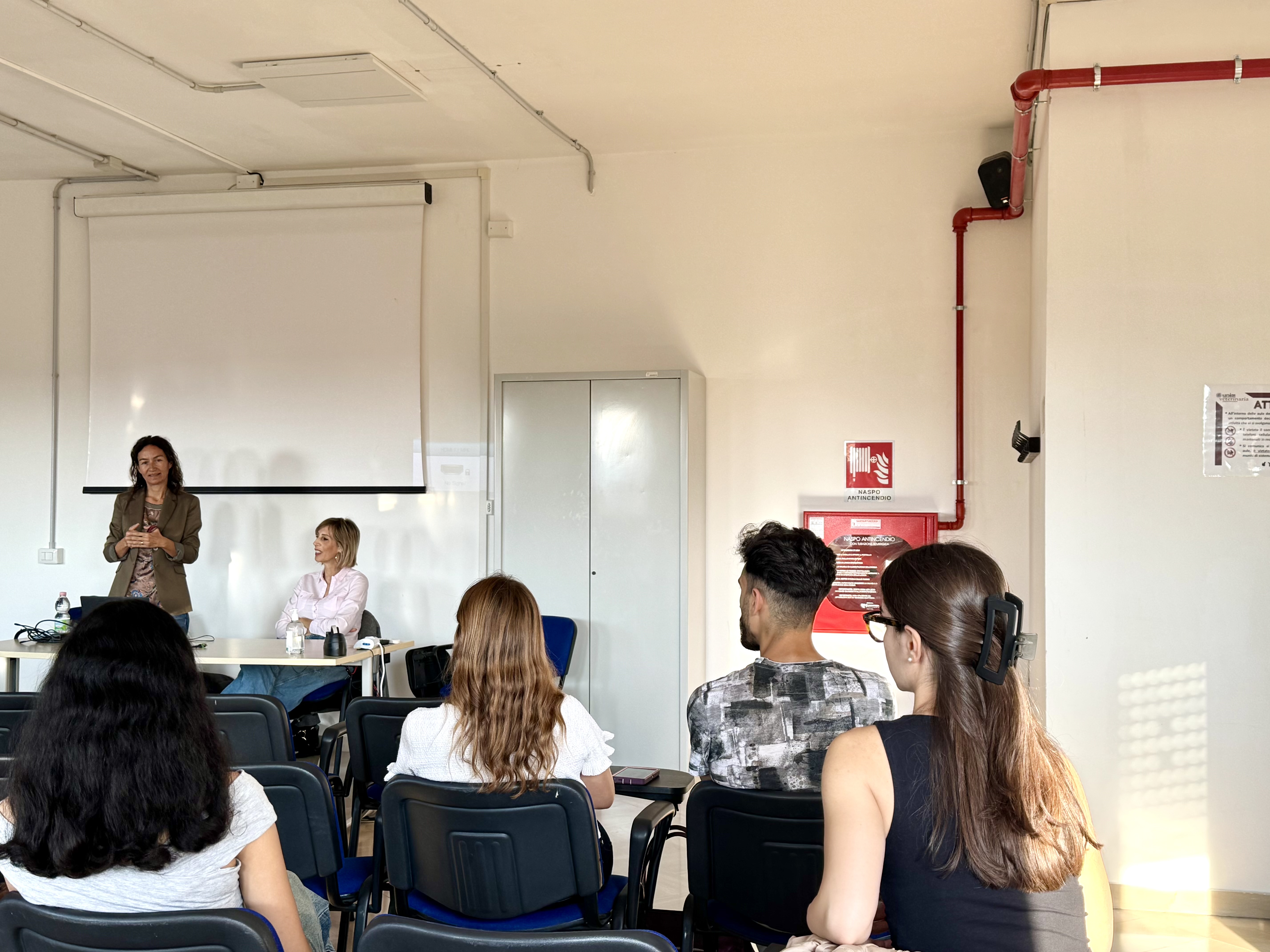 Studentesse e studenti ascoltano la docente in piedi accanto alla cattedra durante il Welcome Day di Biotechnology for Human and Animal Health.