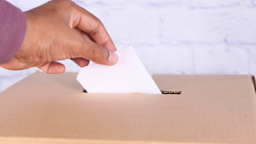 Una mano inserisce una scheda bianca in un’urna di cartone, immagine simbolica di una votazione elettorale