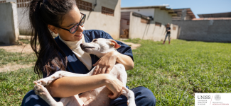 Studentessa di veterinaria accarezza un cane in un'area verde del canile universitario, logo dell'Università di Sassari e del Dipartimento di Medicina Veterinaria in basso a destra.